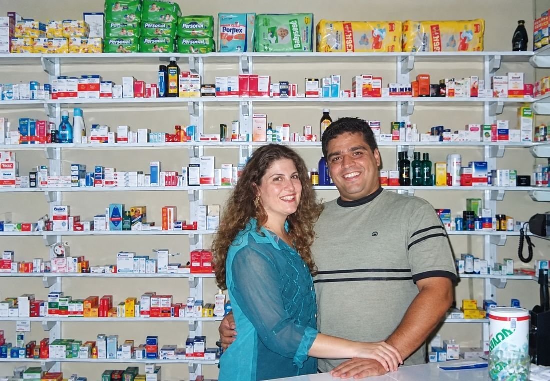 Rodrigo e Aline, donos da D+ Drogaria Popular, sorrindo atrás do balcão da farmácia, com prateleiras de medicamentos ao fundo